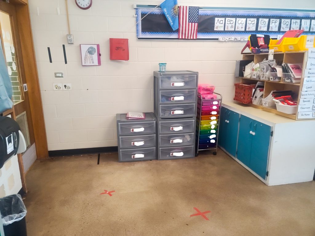 View of plastic drawers with student name label, a rainbow cart, and a wooden shelf filled with adapted books.