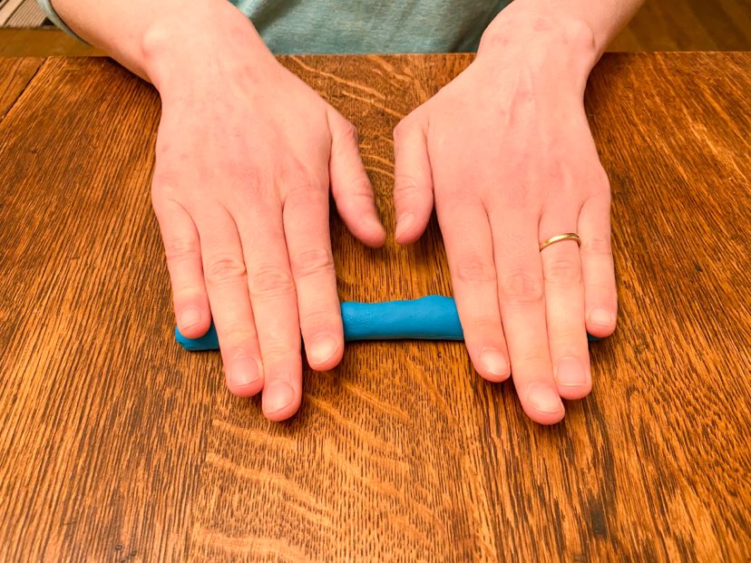 A demonstration of rolling playdough out into a 'noodle'. the author has two hand rolling playdough on a tabletop 