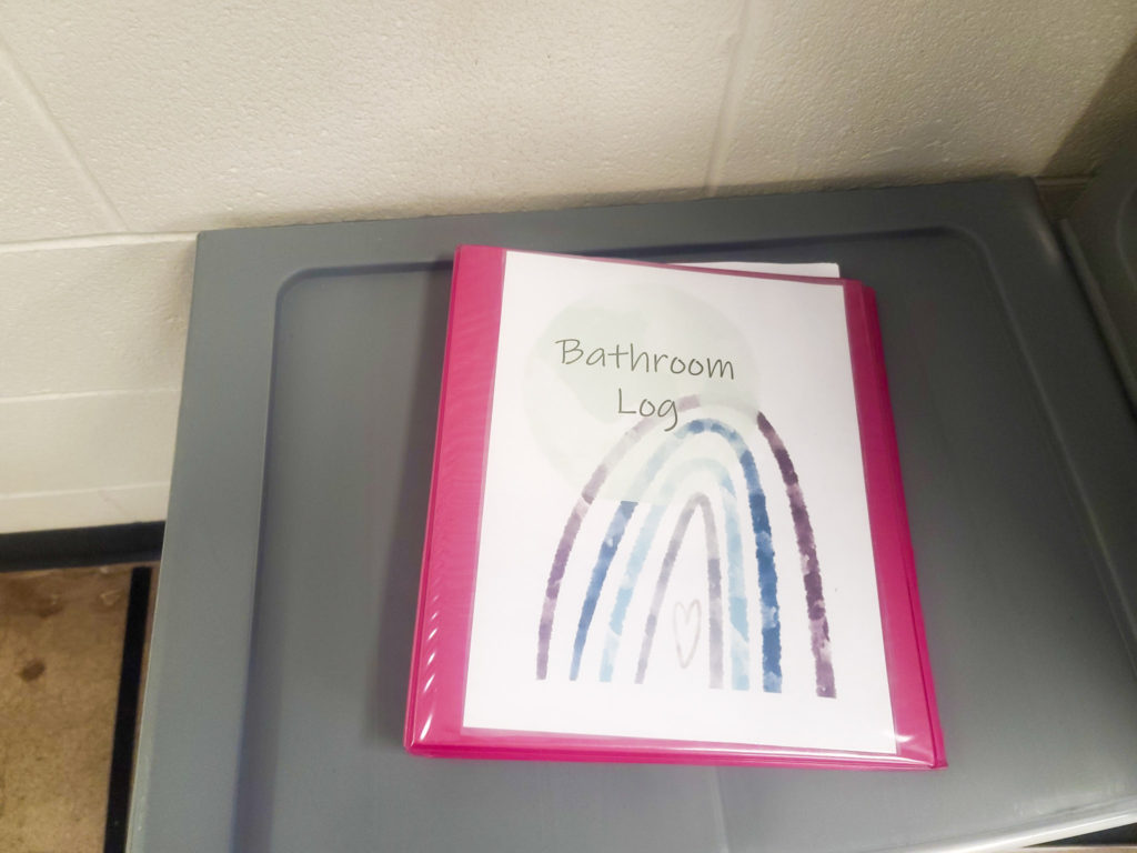 Image of a pink binder on a grey shelf. The binder says bathroom log.