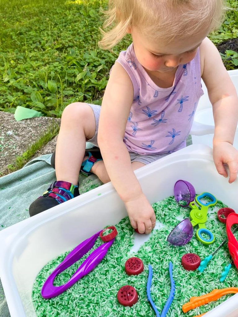 toddler playing with a sensory bin with green rice and different tongs 