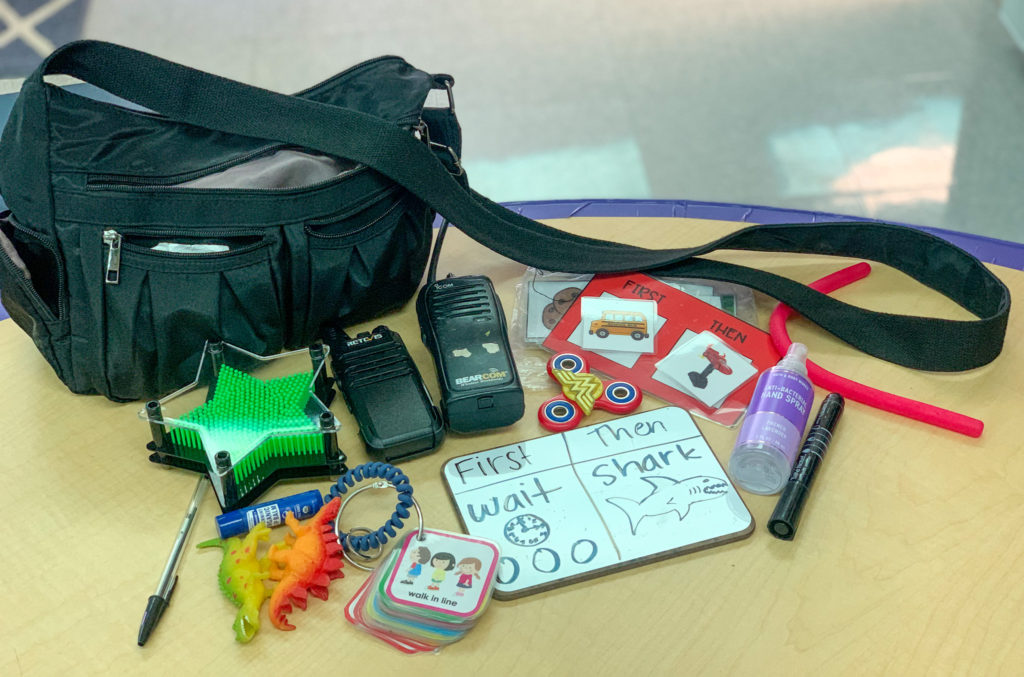 This is a photo of the contents of my teacher bag.  There is a pen, star reinforcer, two colorful dinosaurs, visuals key ring, mini white board, two walkie talkies, a first/then board, a white board marker, some hand sanitizer, a noodle fidget, and a black bag. 