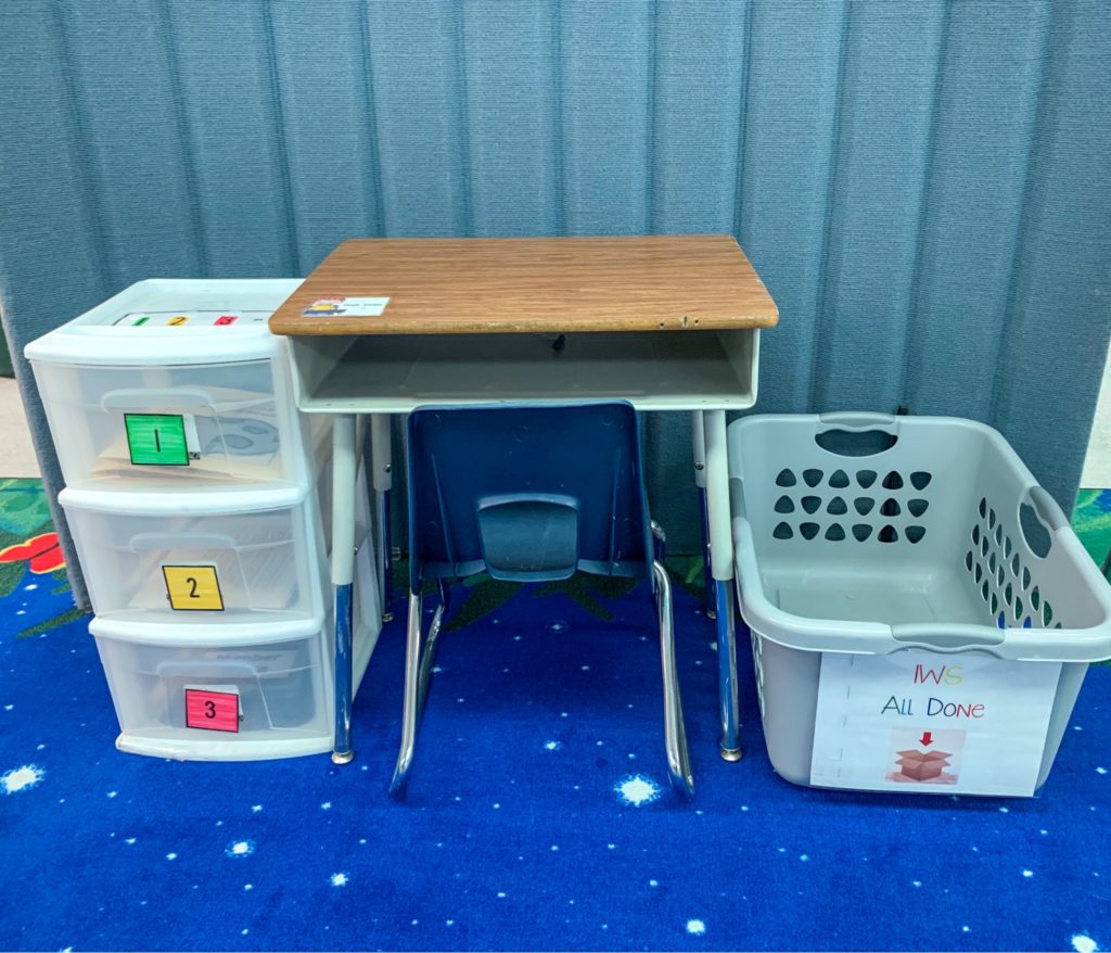 This is a photo of a student desk with a three-drawer bin and a laundry basket next to it.  This is used as a structured work system in special education classrooms. 