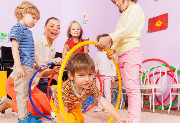 preschool children and teacher holding hoops up for a child to crawl through as he crawls out of a pop up floor tunnel