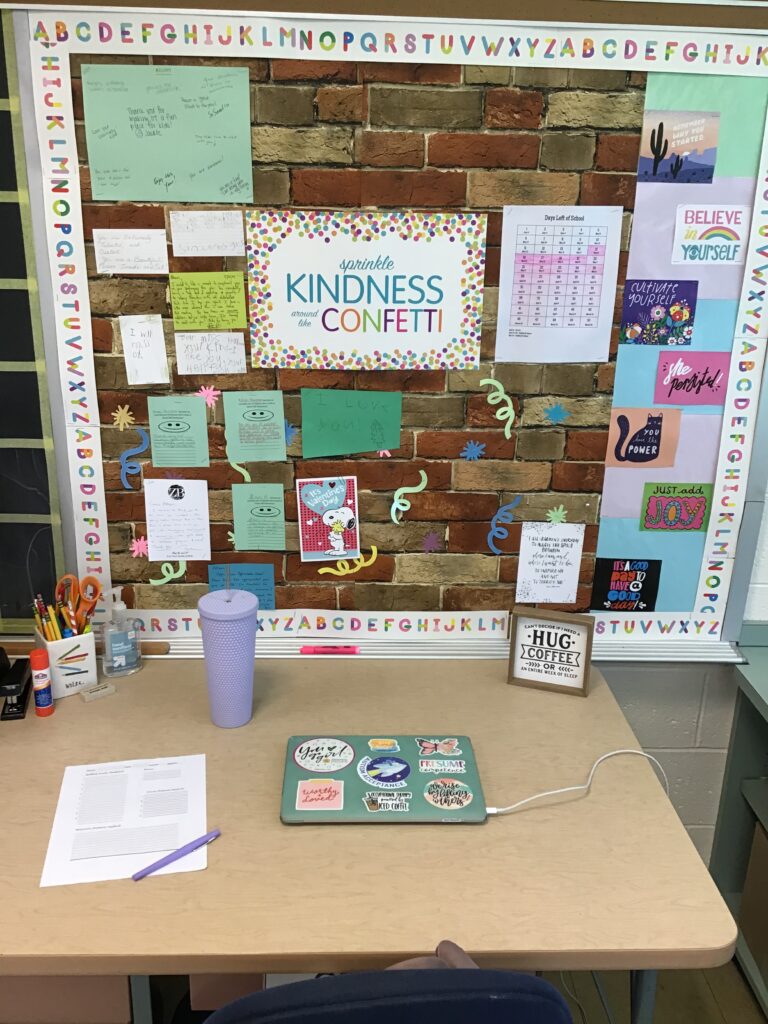 picture of my desk with laptop, stapler, pencil holder, hand sanitizer, purple cup with straw, and adapted spelling test paper.  The bulletin board behind has encouraging quotes and notes from teachers, administrators and students surrounding a sign reading "Sprinkle Kindness Around Like Confetti". 