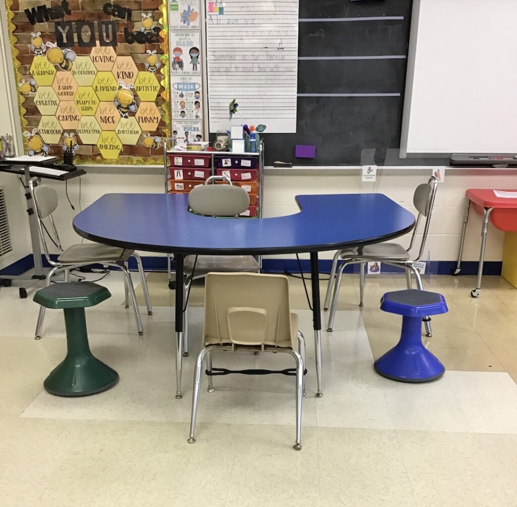 a blue horseshoe-shaped table in my occupational therapy classroom with adapted seating options including wobble stools and bouncy bands as the chairs.  A rainbow drawer cart, chalkboard and bulletin board with beehive design are behind the table.