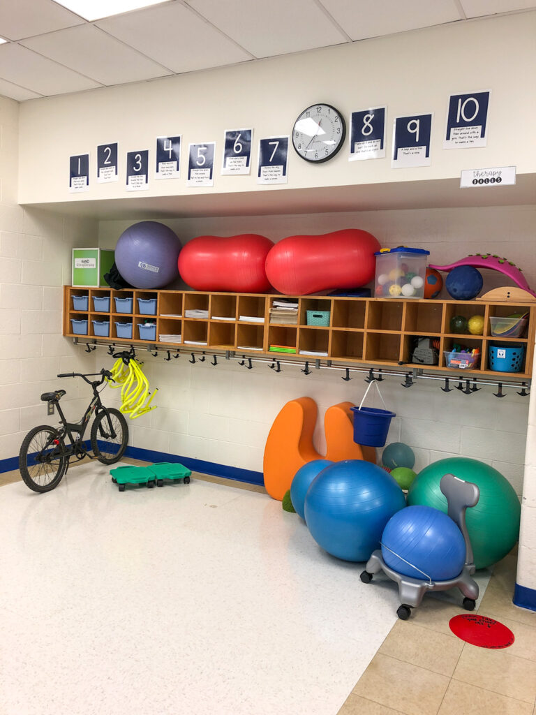 the cubby area of the occupational therapy room with therapy balls stored on top, bins, paper and smaller items in the cubbies and a bike, scooters, adapted seating, and therapy balls under the backpack hooks.
