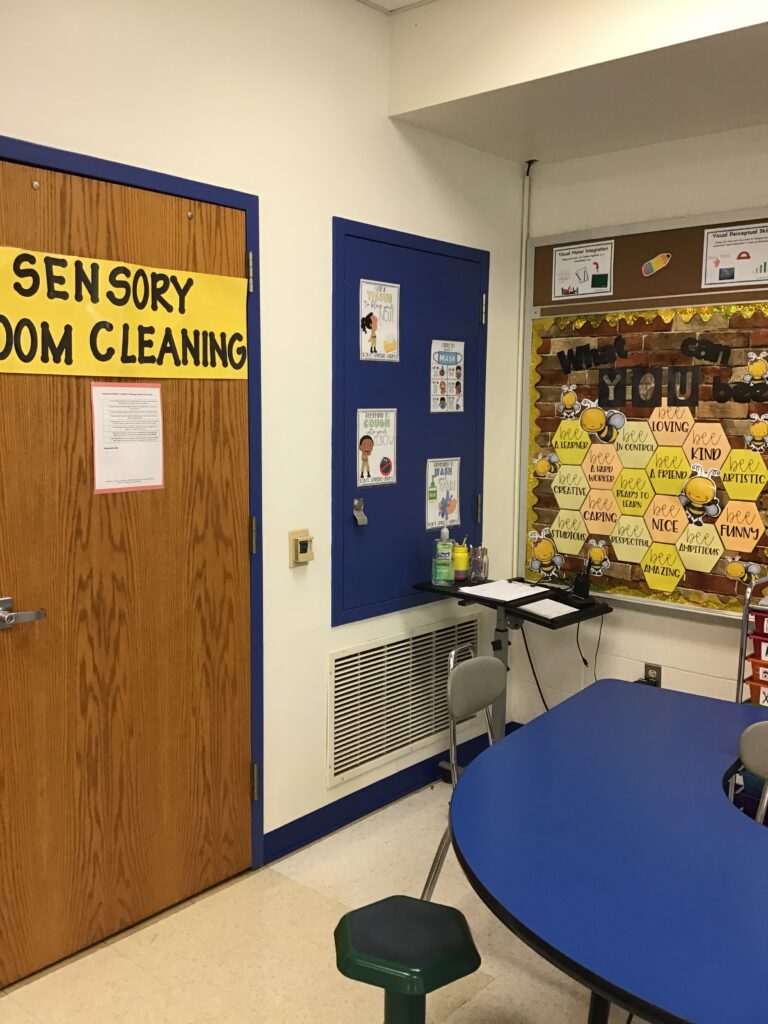 next to the horseshoe-shaped table is a large yellow sign reading "sensory room cleaning" with a checklist below.  In the corner, there is a small table with a sign in sheet and a walkie talkie.