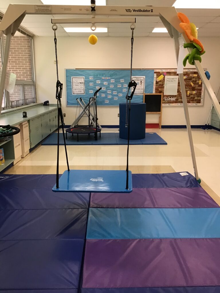 Tumbleform platform swing on an independent frame with mats underneath in the occupational therapy classroom.  A mat with elliptical, trampoline and 3 foot tunnel behind