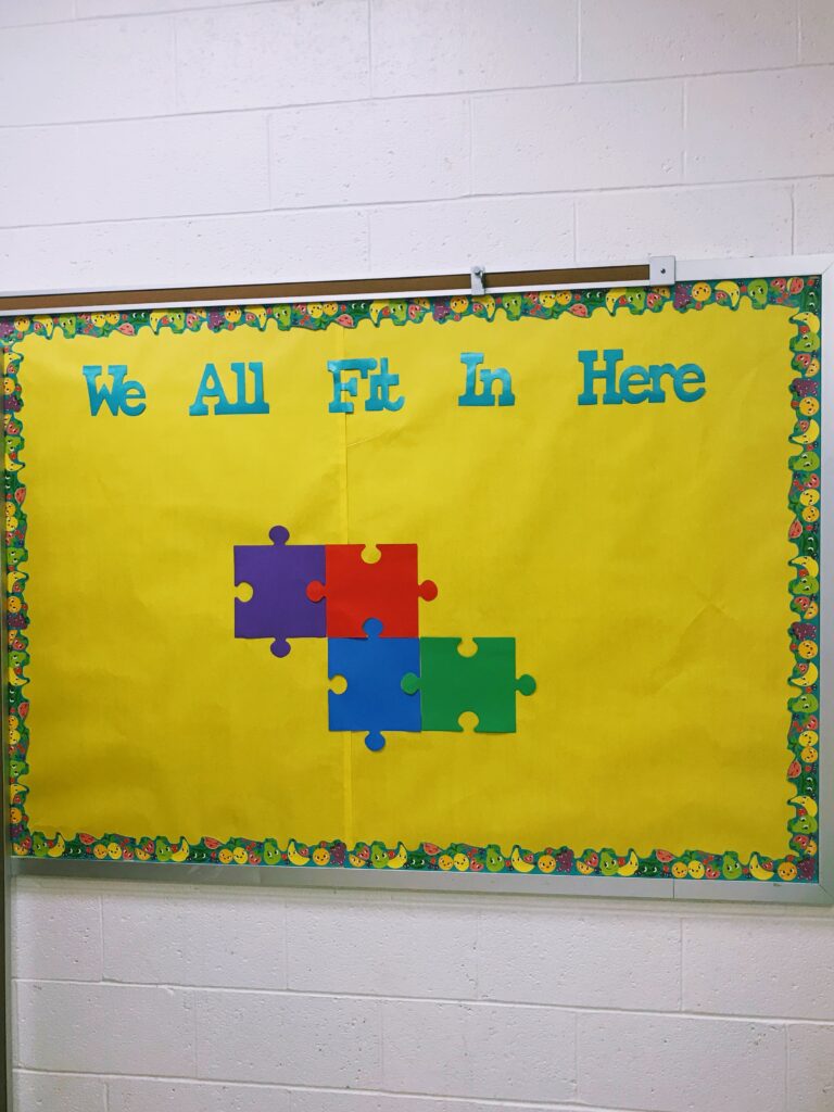bulletin board that says "we all fit in here" with a yellow background and puzzles pieces