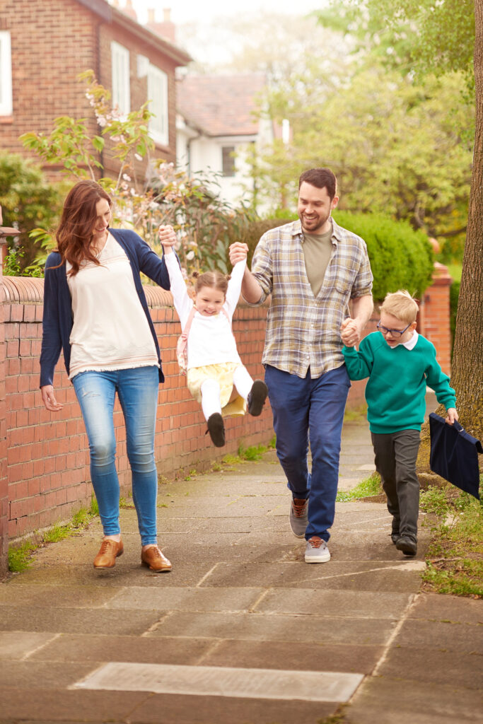 family of four walks down the sidewalk holding hands and swinging the daughter in the air