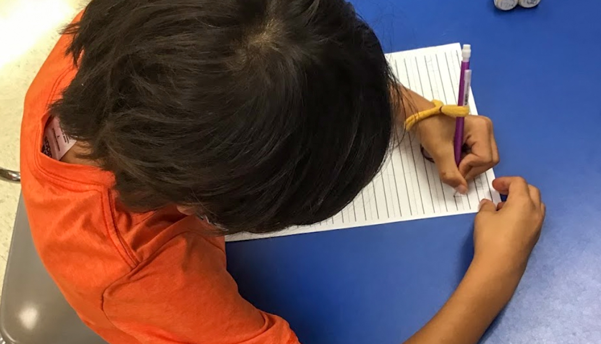 student using a handiwriter pencil grip while writing on three lined paper at a blue table. 