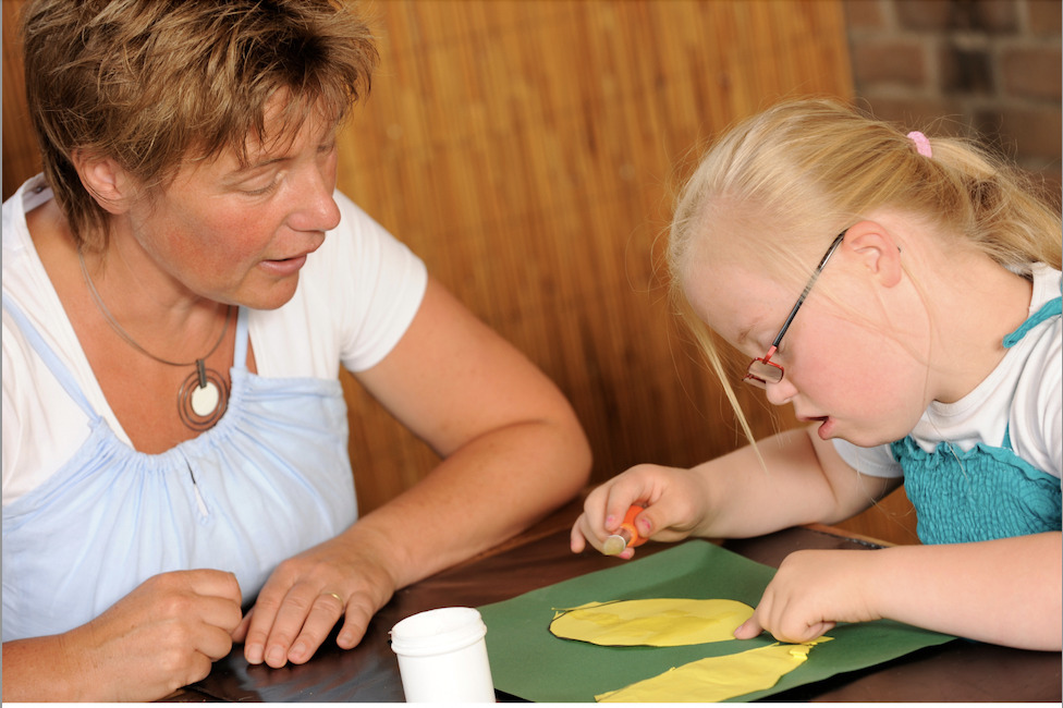 child with Down syndrome works on a craft with the support of an adult who is not using hand over hand assistance, but rather verbal supports