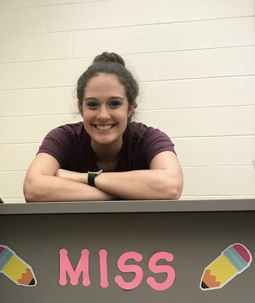 author is leaning on her desk with arms crossed smiling. Pencil images and the beginning part of her name "Miss" is shown on the front of the desk