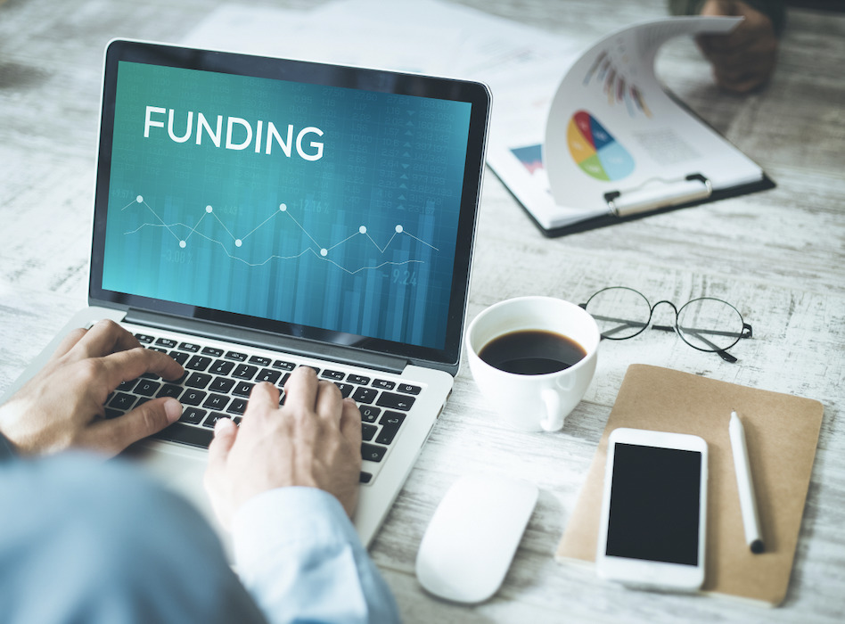 person working on a laptop with the word "funding" on the screen and coffee, cell phone, eyeglasses, pen and paper, etc. surrounding the computer on the desk