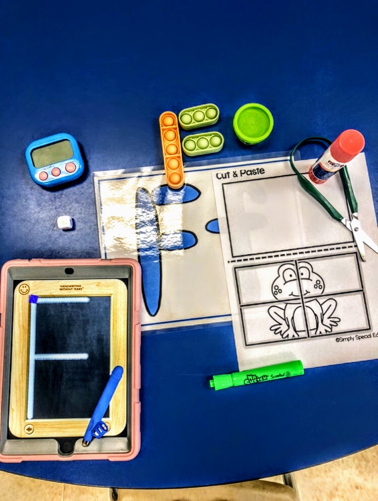 a variety of occupational therapy materials on a blue table including a timer, pop-it fidgets forming the letter F, Handwriting Without Tears app on the iPad with Write Right stylus, play dough, adapted scissors, a glue stick, Simply Special Ed's cut and paste resource with a frog and a Mr. Sketch scented marker