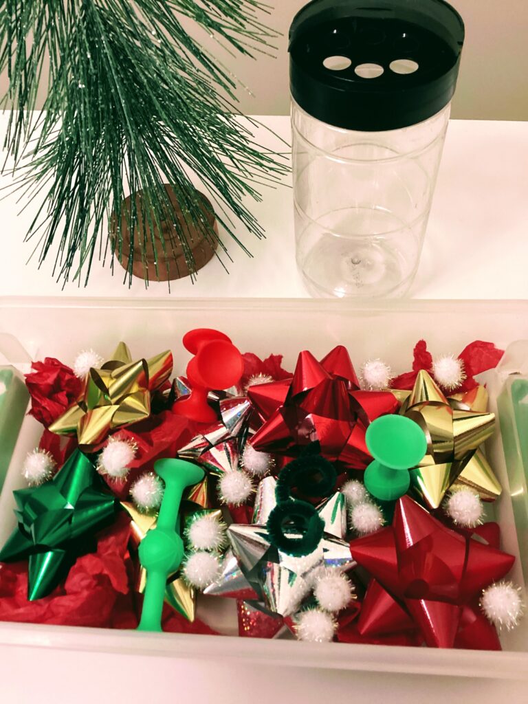 sensory bin pictured above with holiday items including tissue paper for auditory element with an empty container behind it to put pom poms in and a mini Christmas tree for decoration