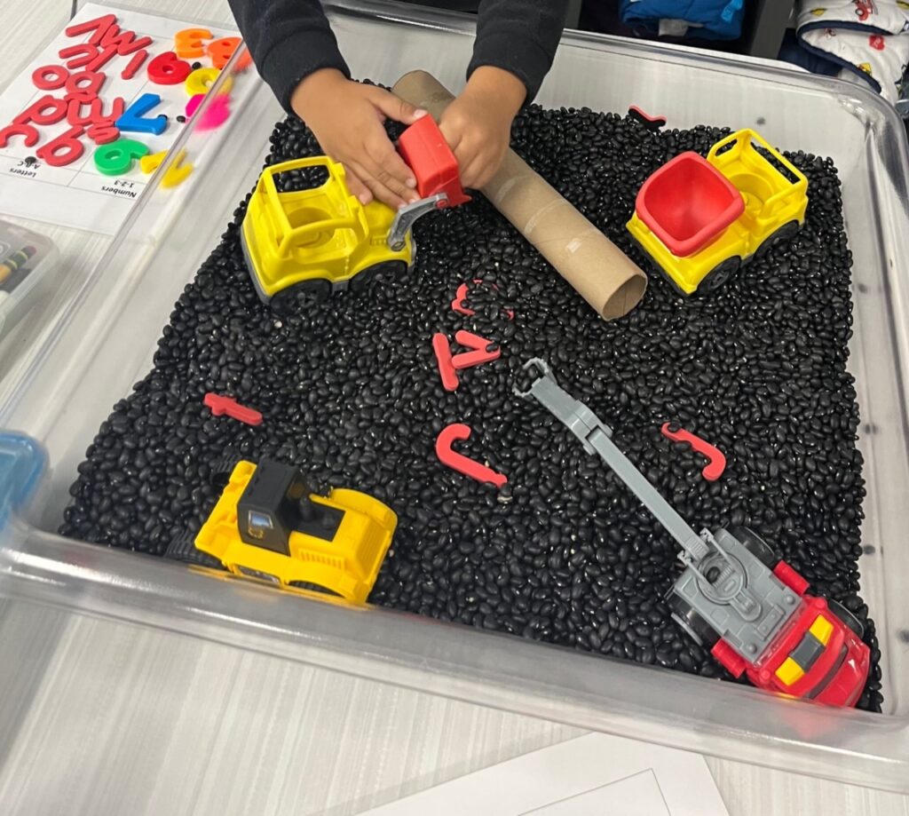 Sensory bin filled with dry black beans , construction themed vehicles and letters. 