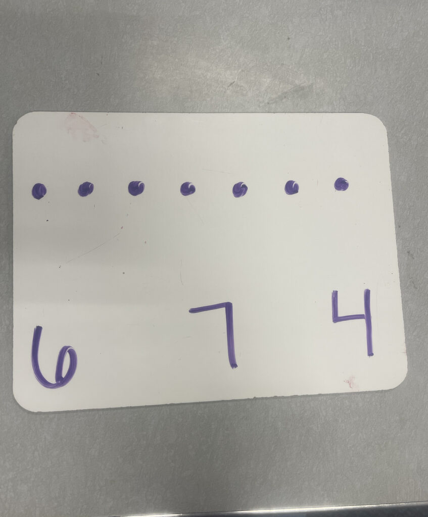 A whiteboard with 7 dots written on it and the numbers 6,7 and 4 written below the dots.