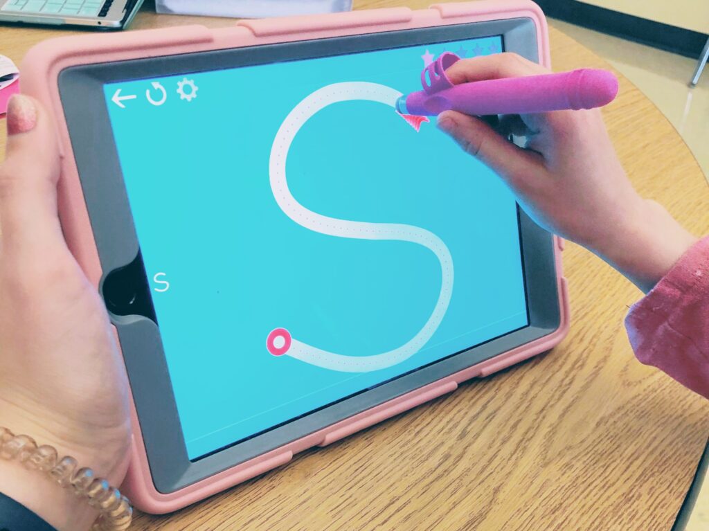 student traces the letter S on an iPad held at a vertical angle by the OT using the Writing Wizard app and a Write Right stylus