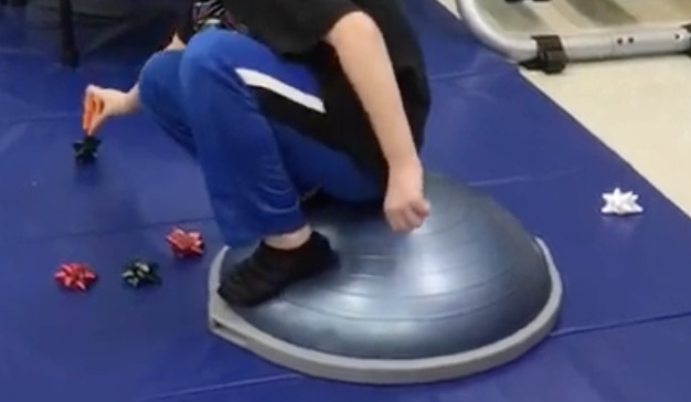 student standing on a boss ball picking up Christmas bows with tweezers