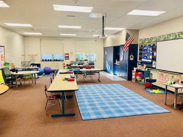 A view of an elementary school classroom. Several student work tables surround a blue rug.
