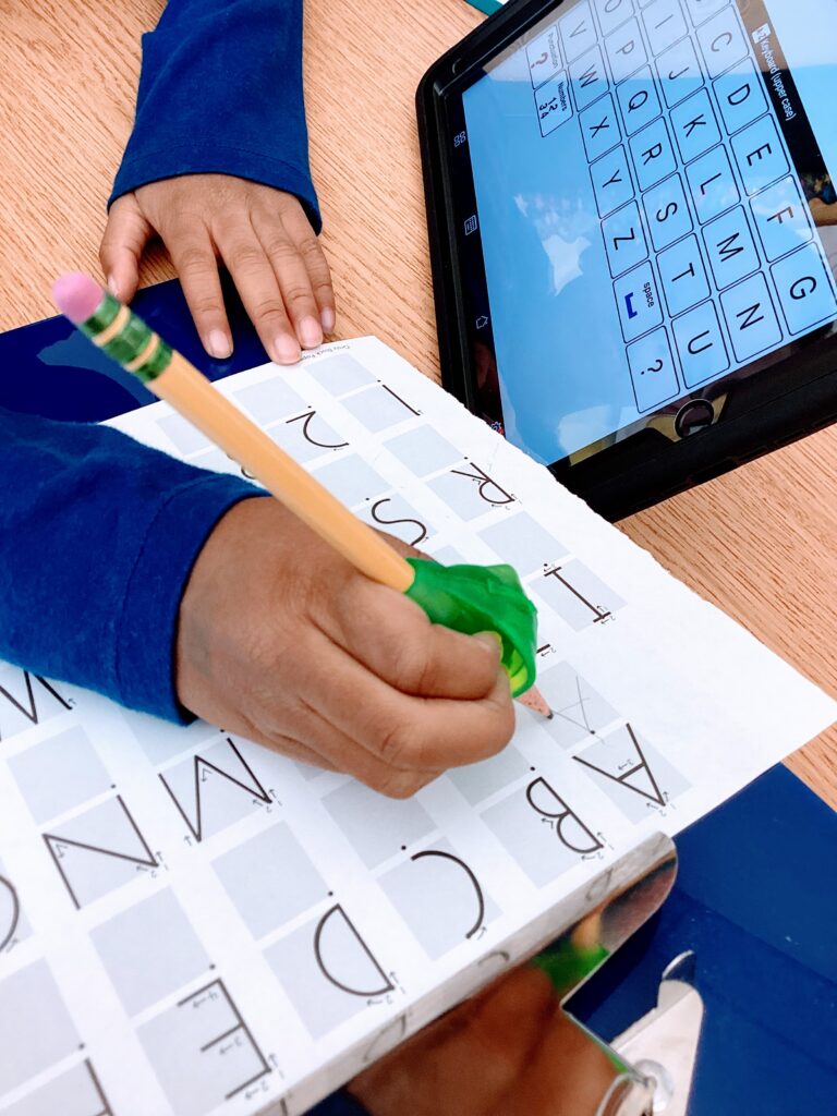 student using a pencil grip and slant board to copy uppercase letters on Handwriting Without Tears paper next to his AAC device