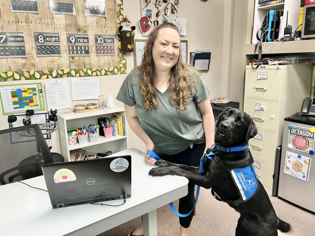 Woman standing at desk in classroom with service dogs front two paws on desk too