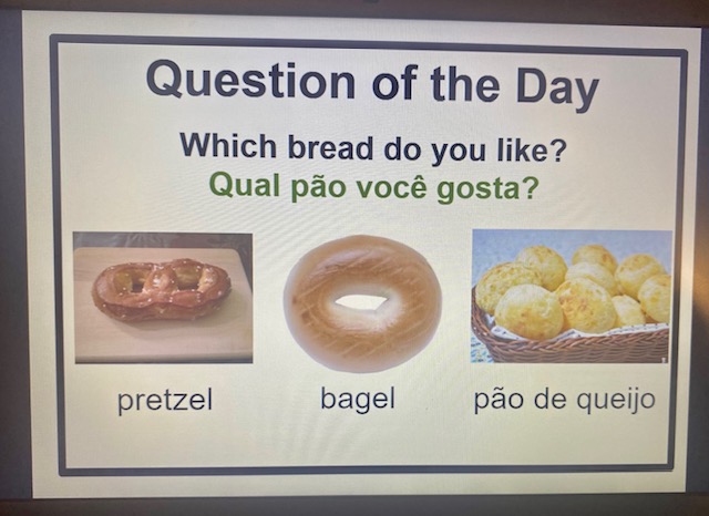 Google Slide showing the morning meeting question of the day in English and Portuguese, with images of pretzel, bagel, and pao de quiejo