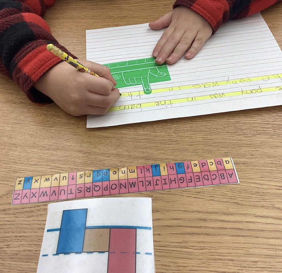 student writing on adapted highlighted paper with a finger spring manipulative and a color coded visual 