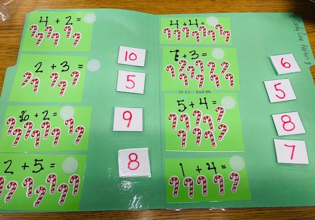 A green file folder activity is open on a table. Inside are addition facts and answers, with candy cane stickers underneath each problem.