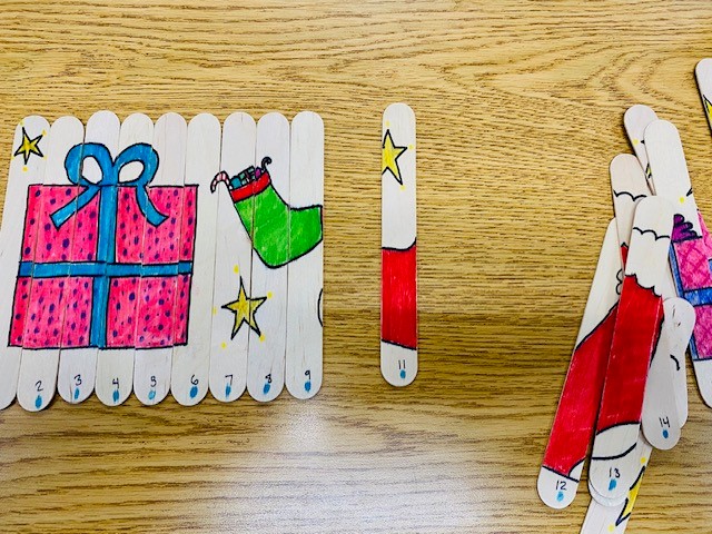 On a wooden table is a set of craft sticks that are decorated with a holiday present and stocking. Some of the pieces are sequenced, and others are in a pile to the side.