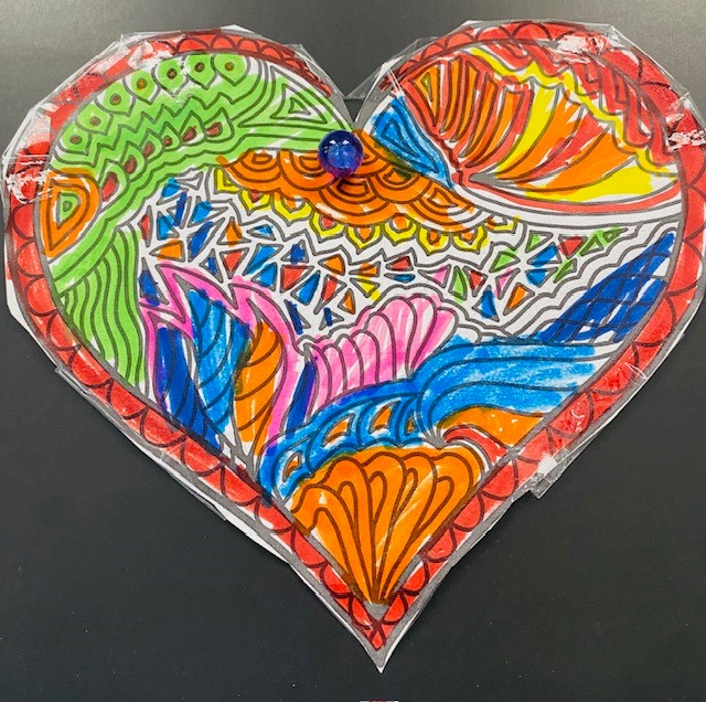 Image of a colorful paper heart on a black background. The heart has different colored designs, and was colored and cut out by a child.