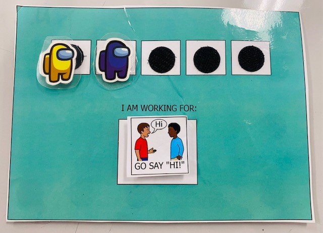 A small blue token board is shown. There are two "Among Us" crewmate tokens and three blank spots. Underneath the tokens, it says "I am working for: Go say hi." There is a small illustration of a boy saying hi.
