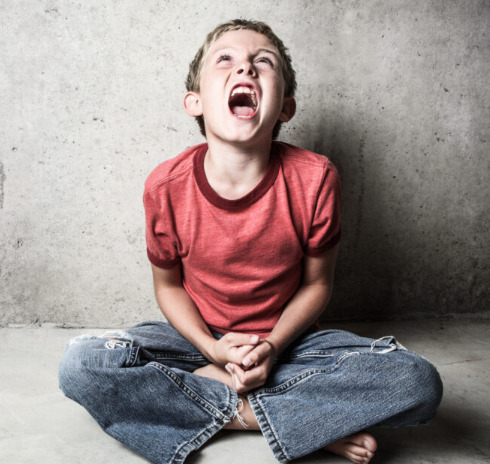 A boy in a red shirt and blue jeans sits on the ground in a gray room. He appears to be yelling.