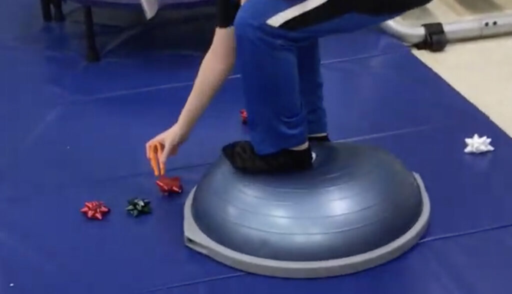 student standing on a boss ball in an obstacle course using tweezers to pick up gift wrap bows