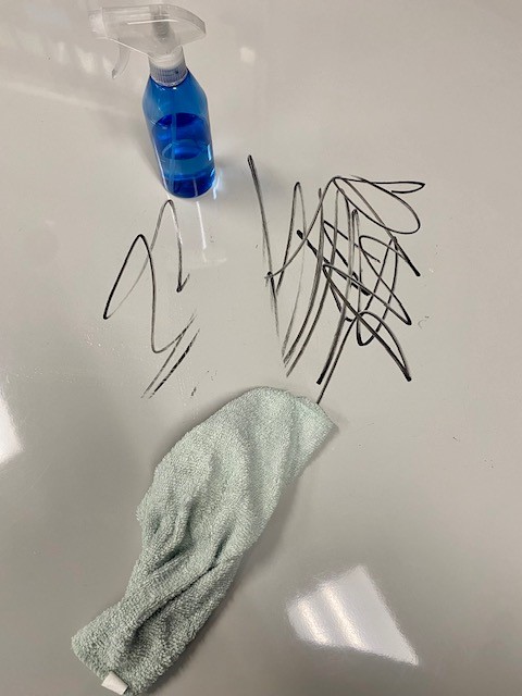 A white dry erase table with black marker marks is shown. There is a blue spray bottle and a light blue towel on the table.