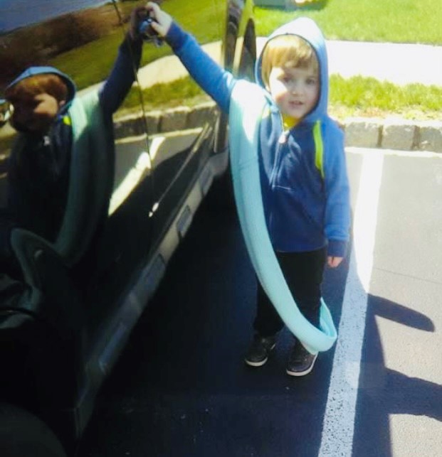 Photo of a young boy standing next to a black car on a sunny day. The boy is wearing long pants, a hoodie with the hood up, and a long, light blue scarf is draped over his body.