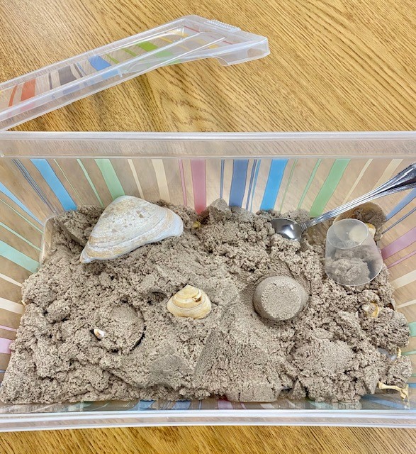 A plastic bin on a wooden table, with tan-colored kinetic sand and seashells in it.