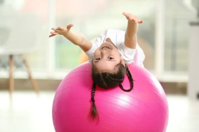 A young girl with brown hair lies on her back on a bright pink yoga ball.