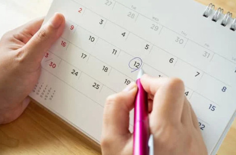 A white calendar page is shown. It is being held in one hand. The other hand is holding a pink pen, circling a date.