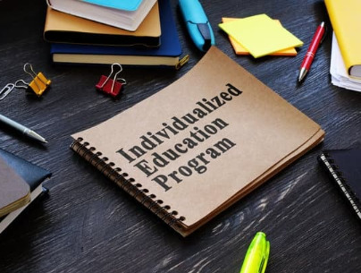 A brown notebook with the words "Individualized Education Program" sits on a wooden table, with various pens and office supplies around it.