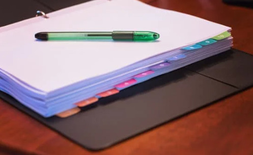 A black binder sits open on a table. A white page is showing, and a green pen sits on top of it.