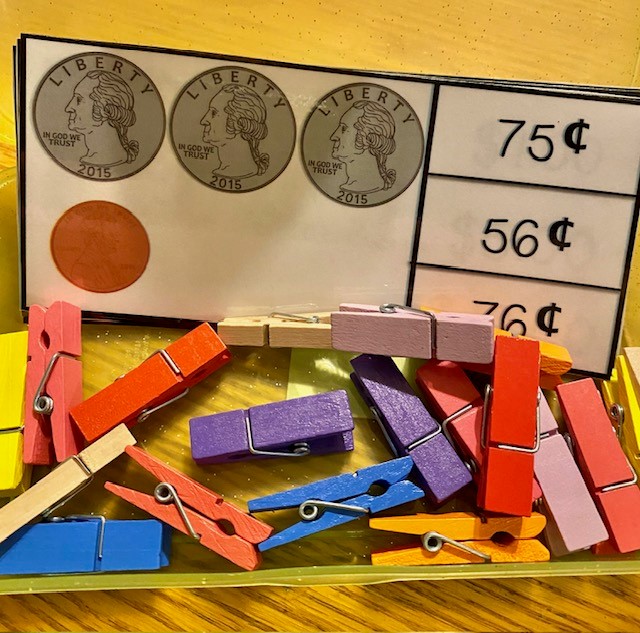 A clear pencil box containing coin-counting task cards, with colorful clothespins.
