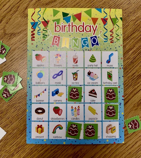 A colorful card that says "birthday bingo" is on a wooden table. The bingo markers are small paper squares with pictures of a chocolate cake. They are on and next to the game board.