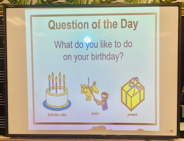 A classroom interactive board is shown, displaying the words "Question of the Day, What do you like to do on your birthday?" There are boardmaker-style images of a birthday cake, pinata, and present.