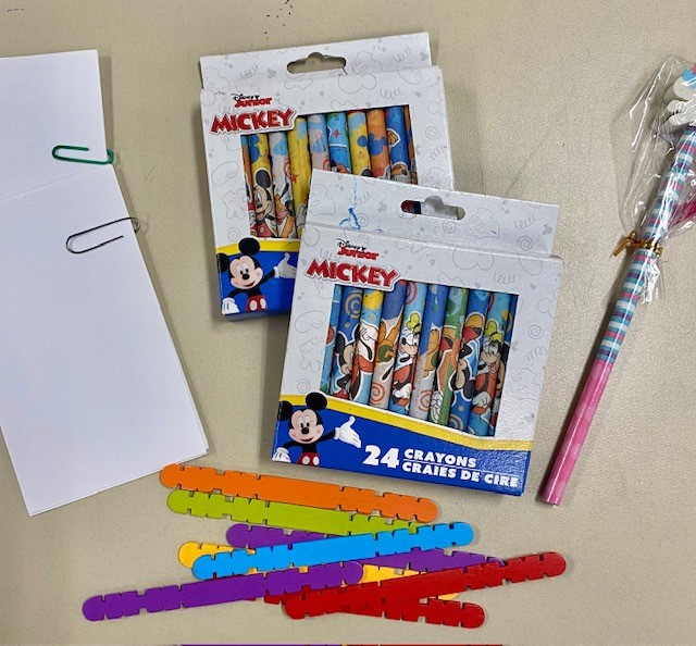 A variety of objects are shown on a table, including white index cards that are paper clipped together; two boxes of crayons; some colorful craft sticks; and a pink and blue striped pencil.