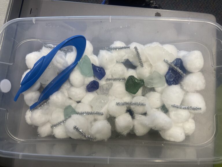 cloudy sensory bin made with cotton balls, pipe cleaners and stones. Picture of the bin includes blue tongs for fine motor