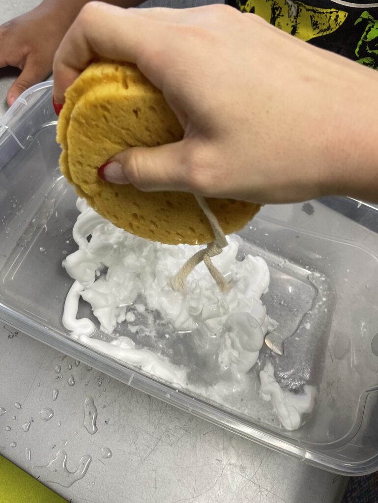 rainy sensory bin including a wet sponge to squeeze as "rain" [hand pictured squeezing sponge] into a bin with shaving cream