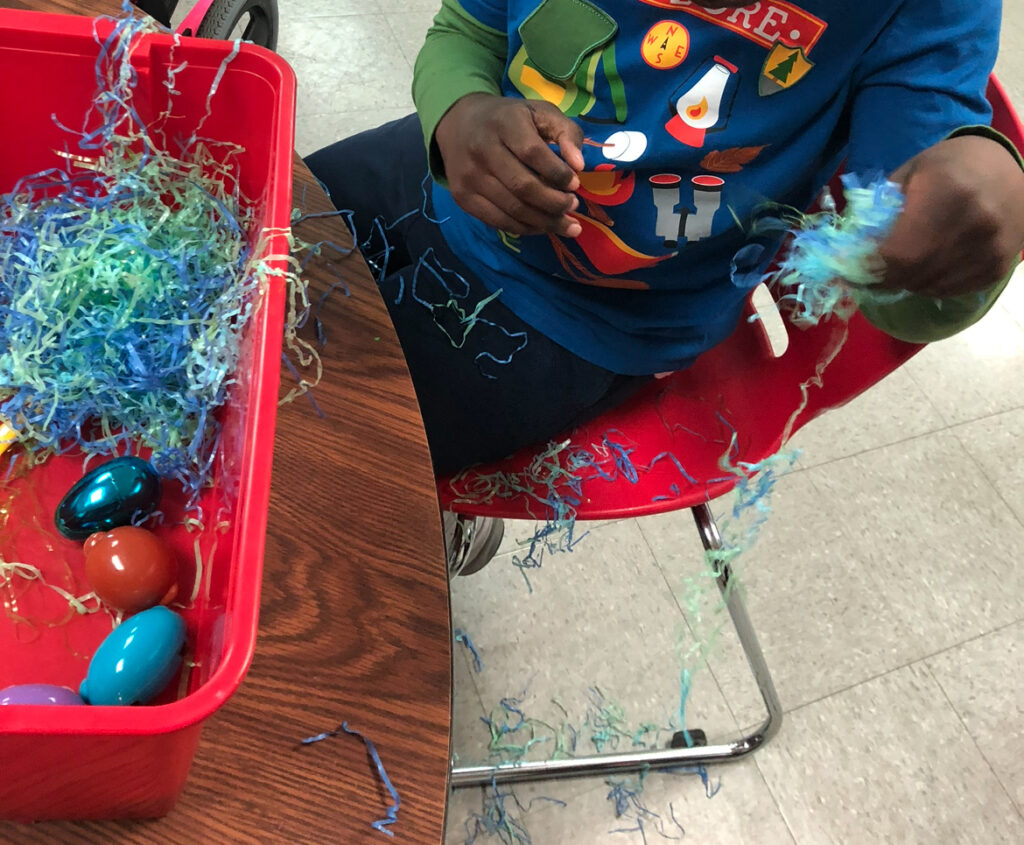 Red bin with Easter grass and Easter eggs being used by a student as a sensory bin. Student sprinkles Easter grass on the floor beside him.