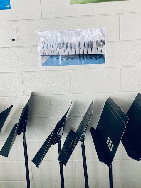 Black music stands are lined up against a wall. Above the stands is a photograph of how the stands are supposed to look.